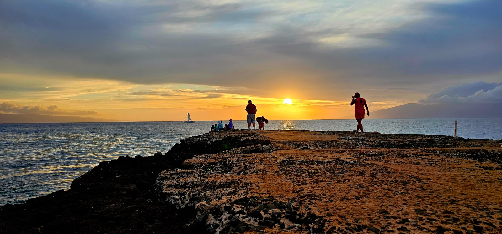 People watching the sun set from a rocky lava point, with a catamaran sailing offshore