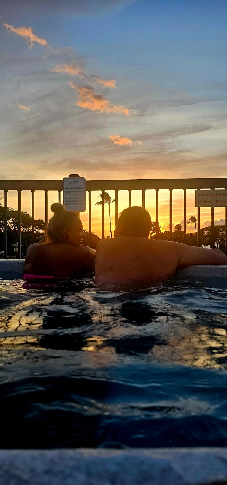 Golden sunset sky over palm trees, seen from a hot tub where two people relax in the warm water